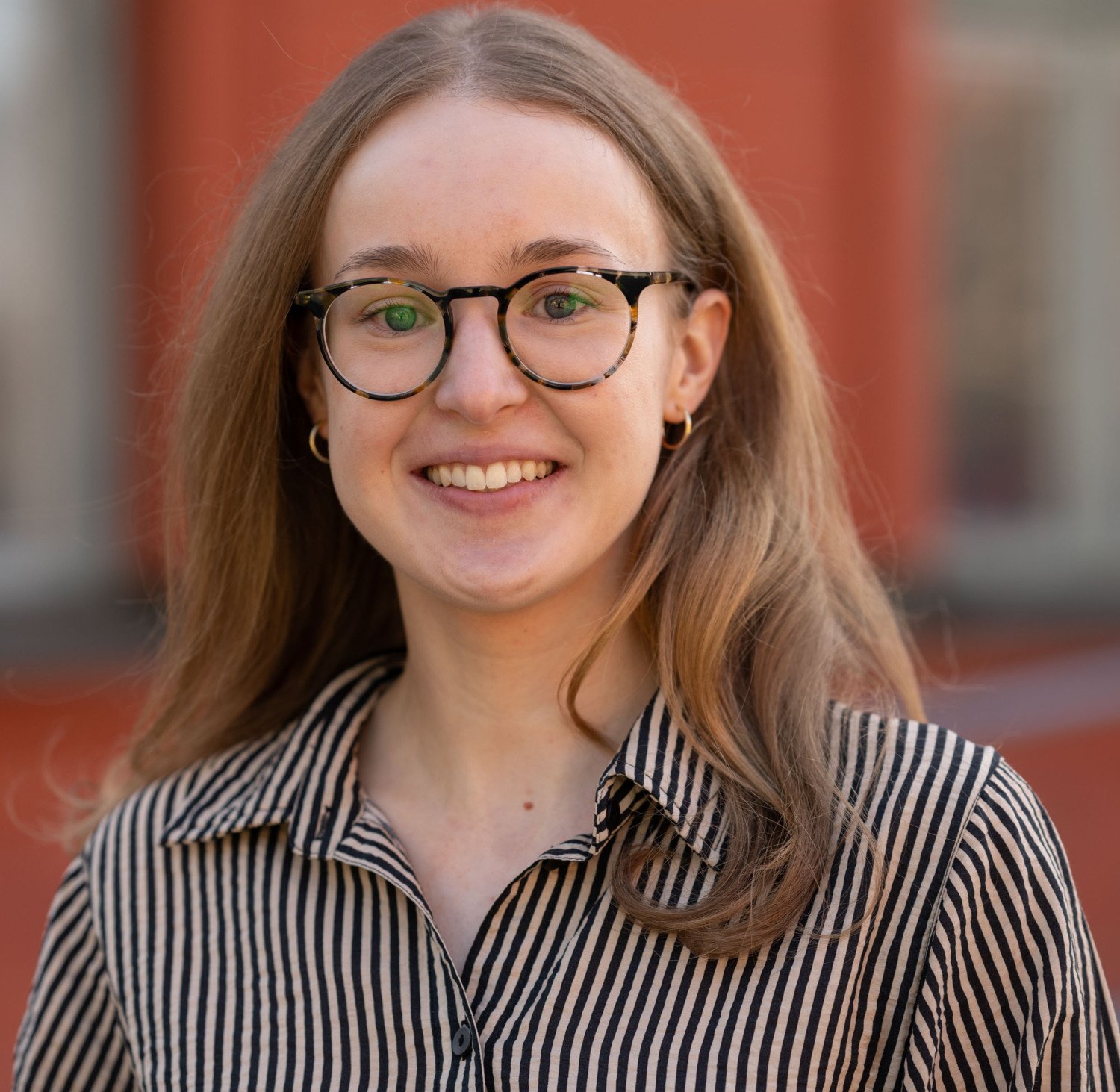 A photo of Theresa Lemke. She is waring a patterned shirt and is standing in front of a red building.