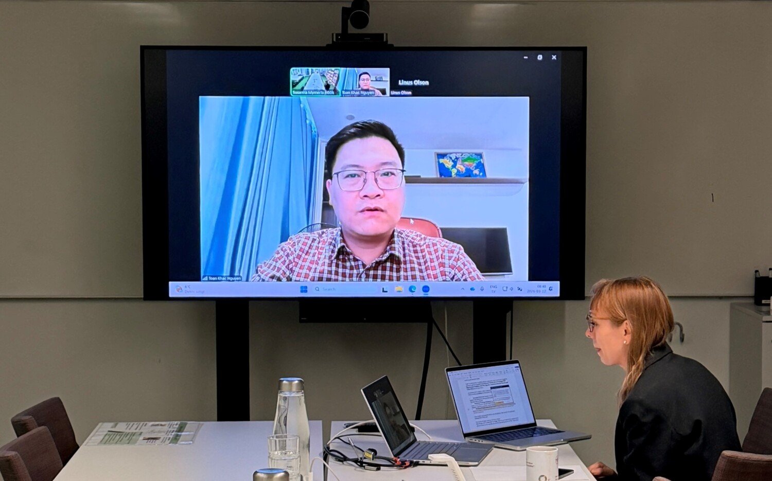 A large screen showing a man, Toàn, presenting his research. Below the screen, a woman is seated at a conference table working on her computer.