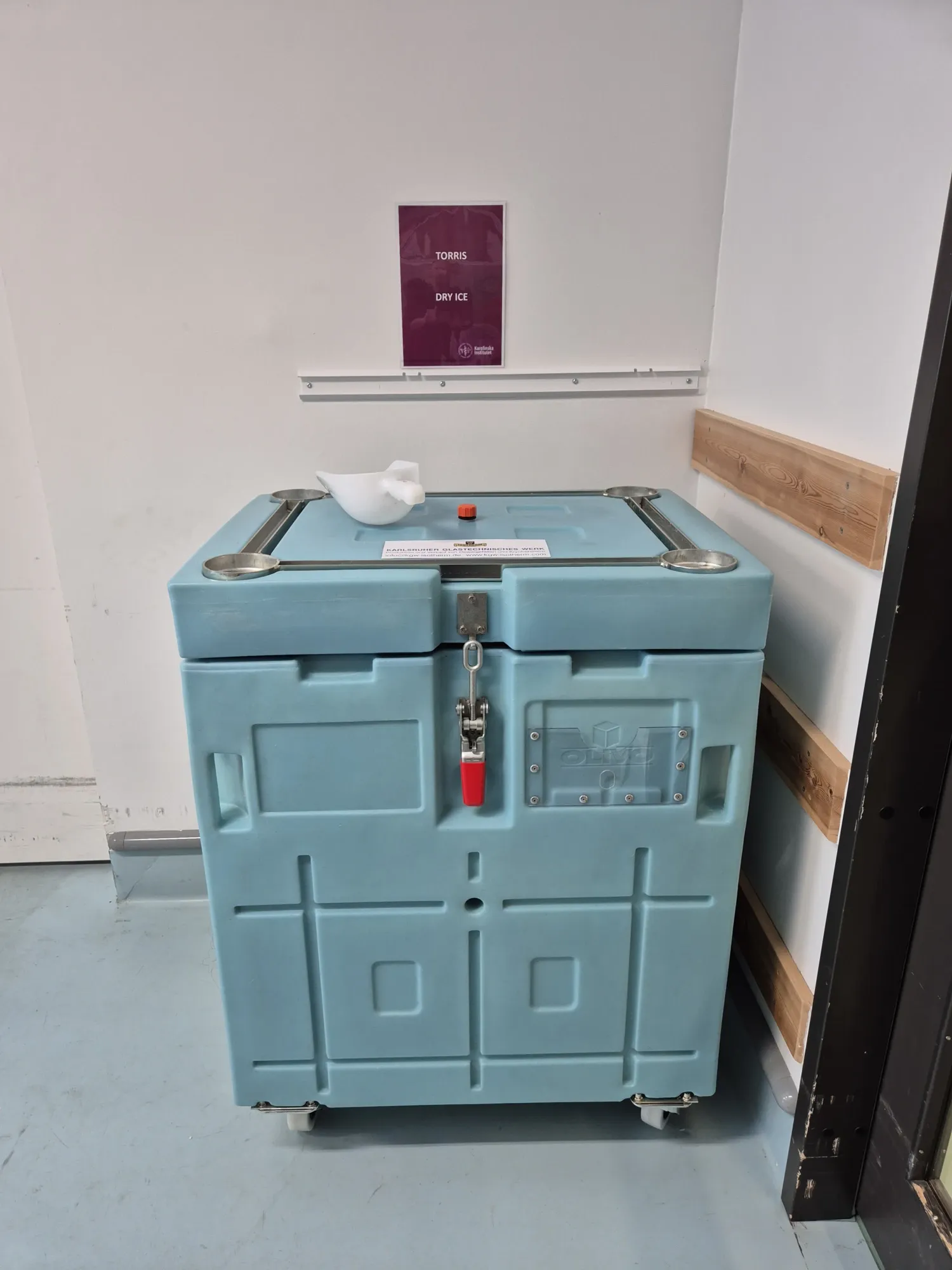 Image of a turquoise dry ice box on wheels standing in a room.