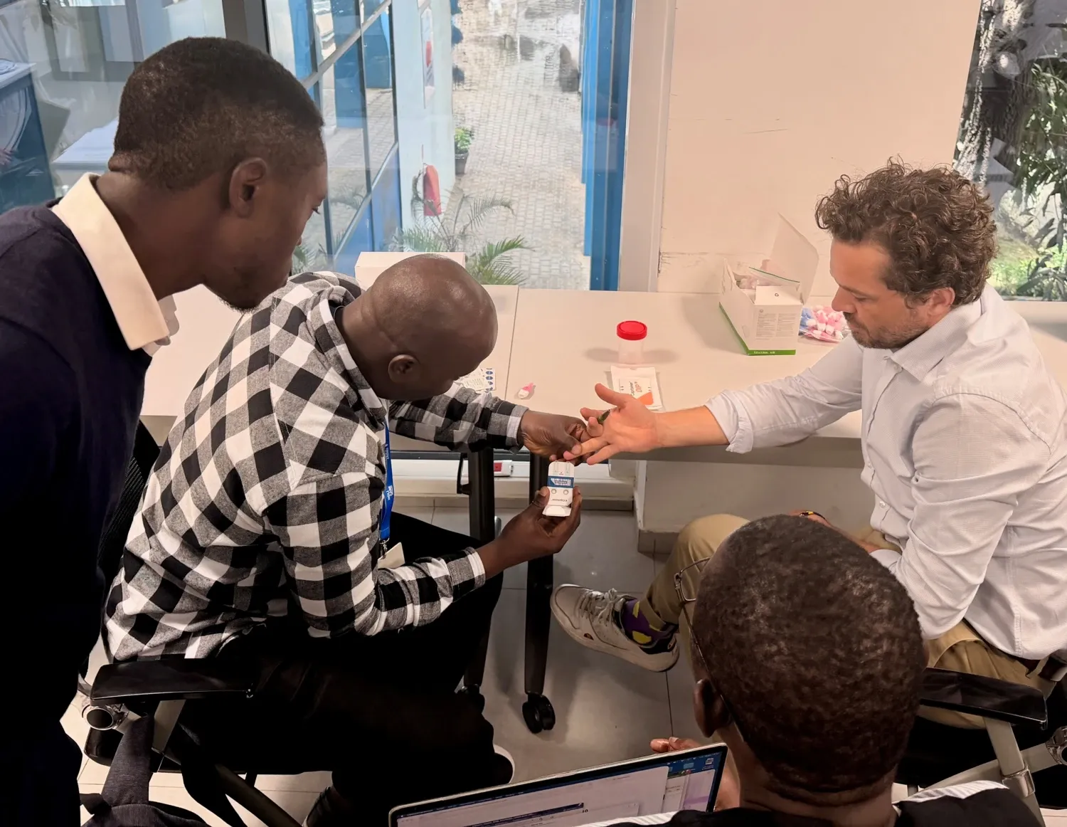 A man is pretending to take a blood sample from another person's finger, while two persons observe.