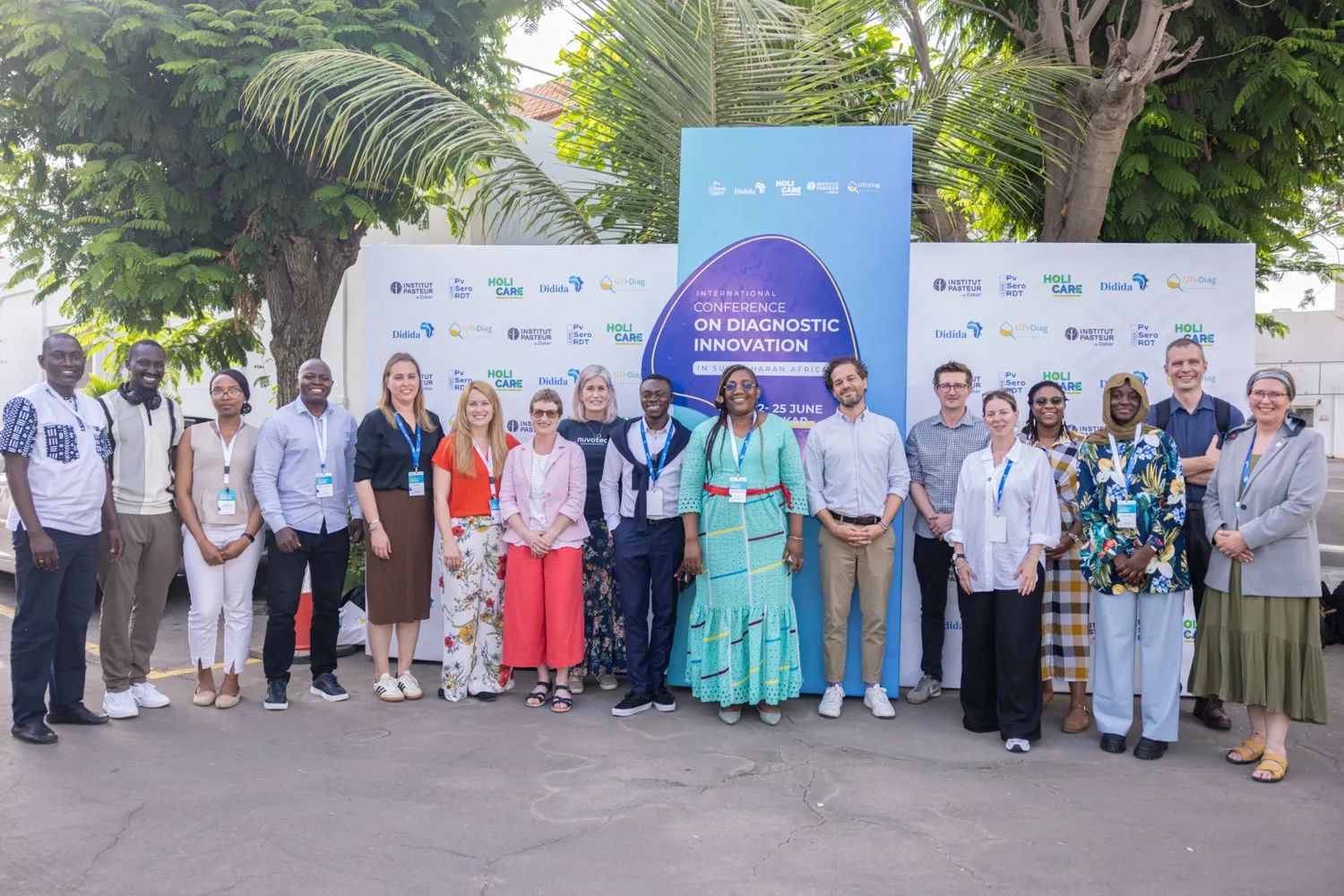 17 persons posing for a group photo, outdoors in front of a big sign with the text International conference on diagnostic innovation.