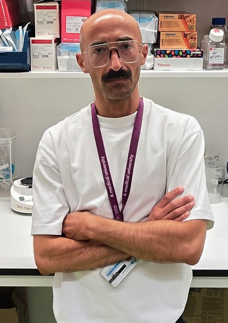 Man in white t-shirt and glasses standing in front of a lab bench.