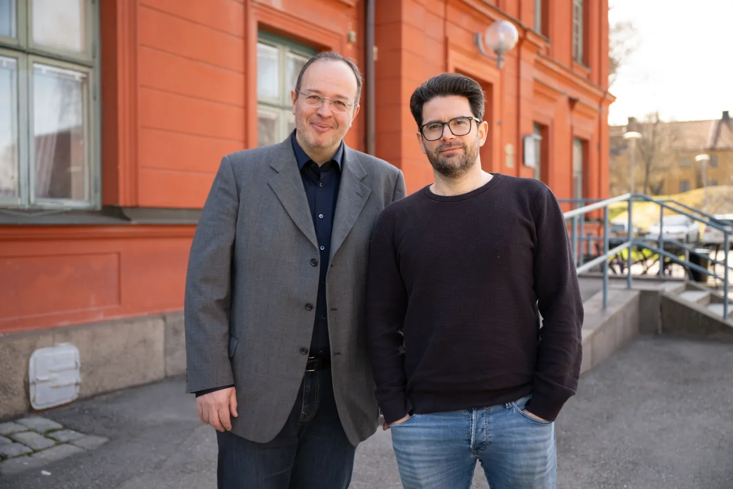 Vladimir Carli, wearing a gray jacket, and Gergö Hadlaczky, wearing a black sweater, standing in front of a red building.