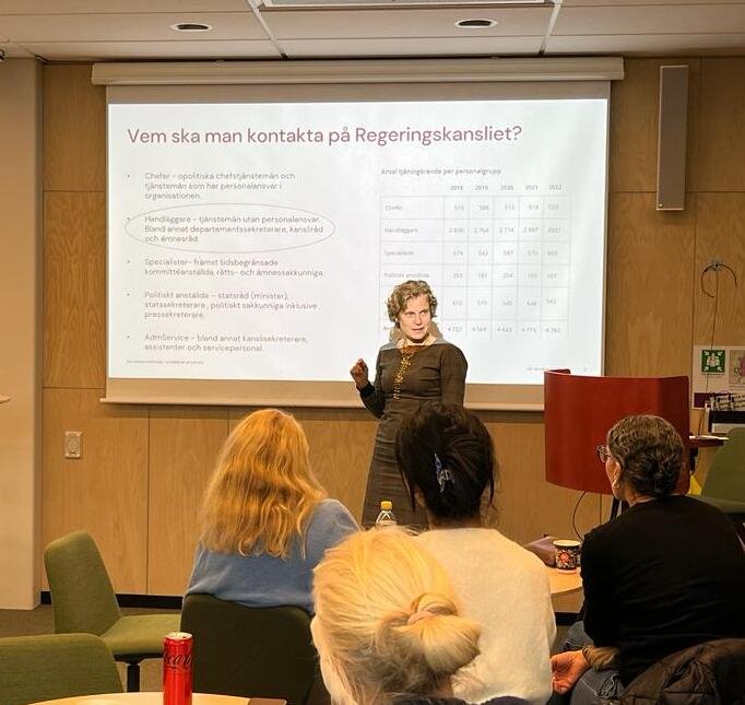 A woman in a dark dress standing in front of a screen showing a powerpoint presentation