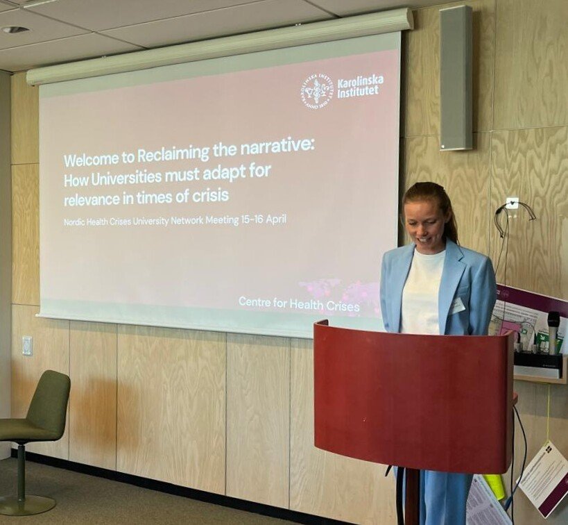 A woman standing in front of a projector screen that says Reclaiming the narrative: How Universities Must Adapt for Relevance in Times of Crisis and the back of a woman sitting in front of it