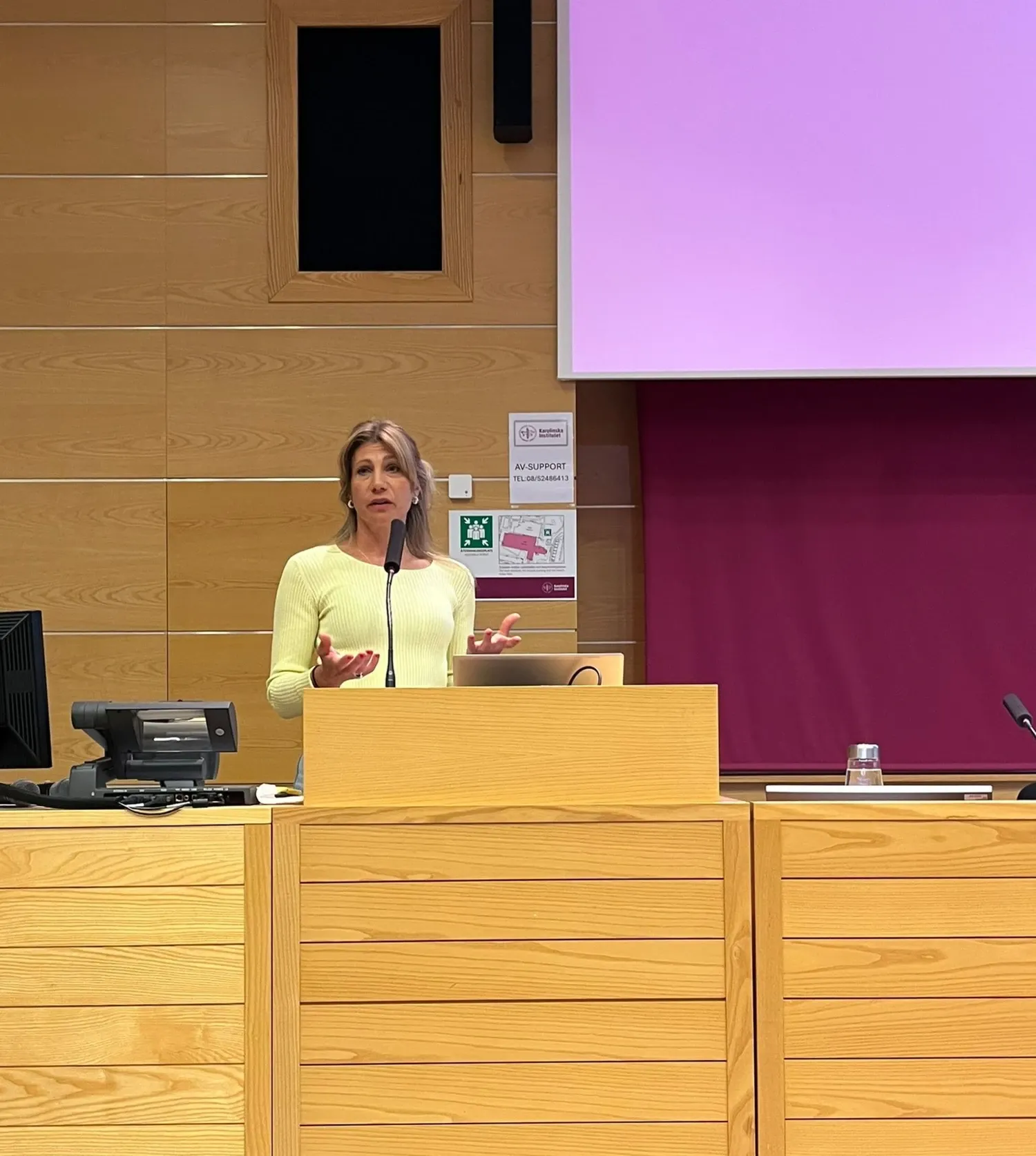 A woman is standing behind a lectern in a lecture theatre; in the background, parts of a screen showing a PowerPoint presentation can be seen