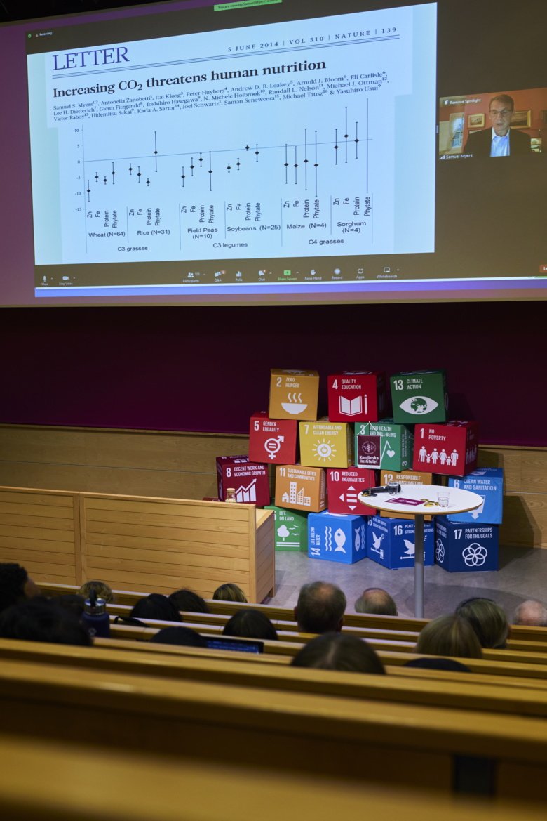 A stage with UN:s Sustainable Development Goals in qubes. A digital presenter shows a power point slide with title "Increasing CO2 threatens human nutrition".