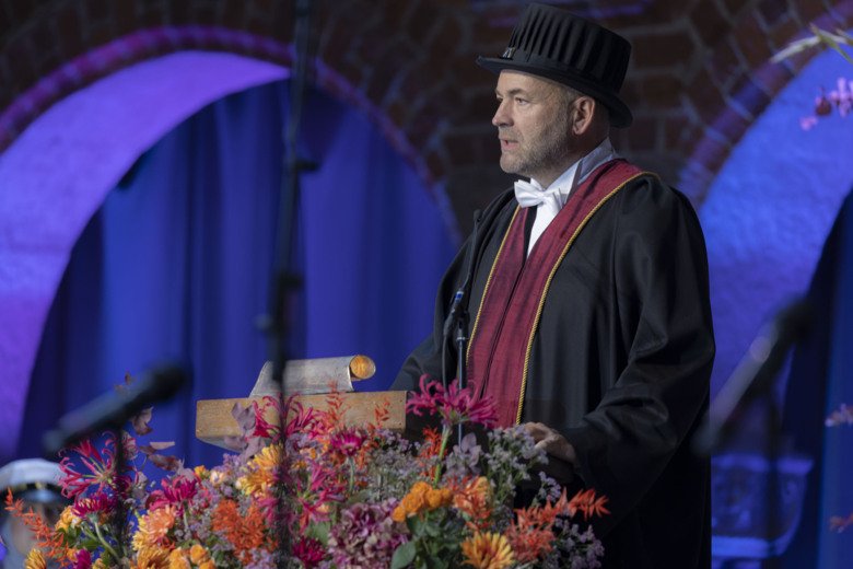 Robert Harris in his doctoral hat, standing on stage facing the crowd.