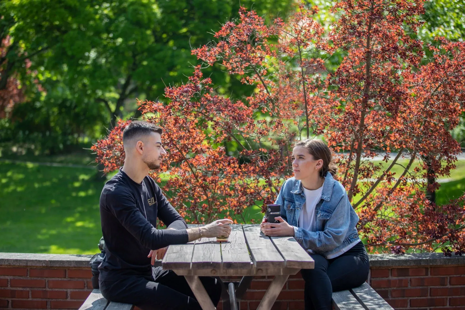 Two students sit at a wooden table at Jöns Jacob's outdoor terrace at Solna campus on a sunny spring day.