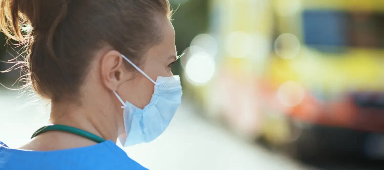 A woman with a face mask, turning to the side, in the background we see the blurred shape of an ambulance
