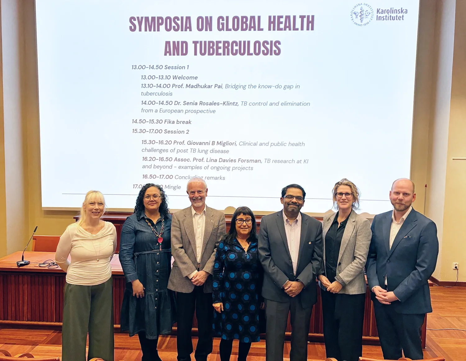 Seven people standing in an auditorium. Behind them, a large screen is displayed