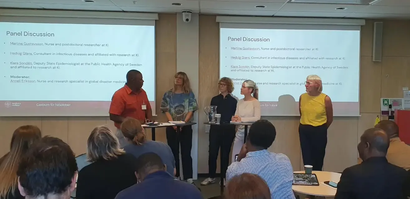 Five people – one man and four women – are standing at a high table, in front of a projector screen displaying their names and titles