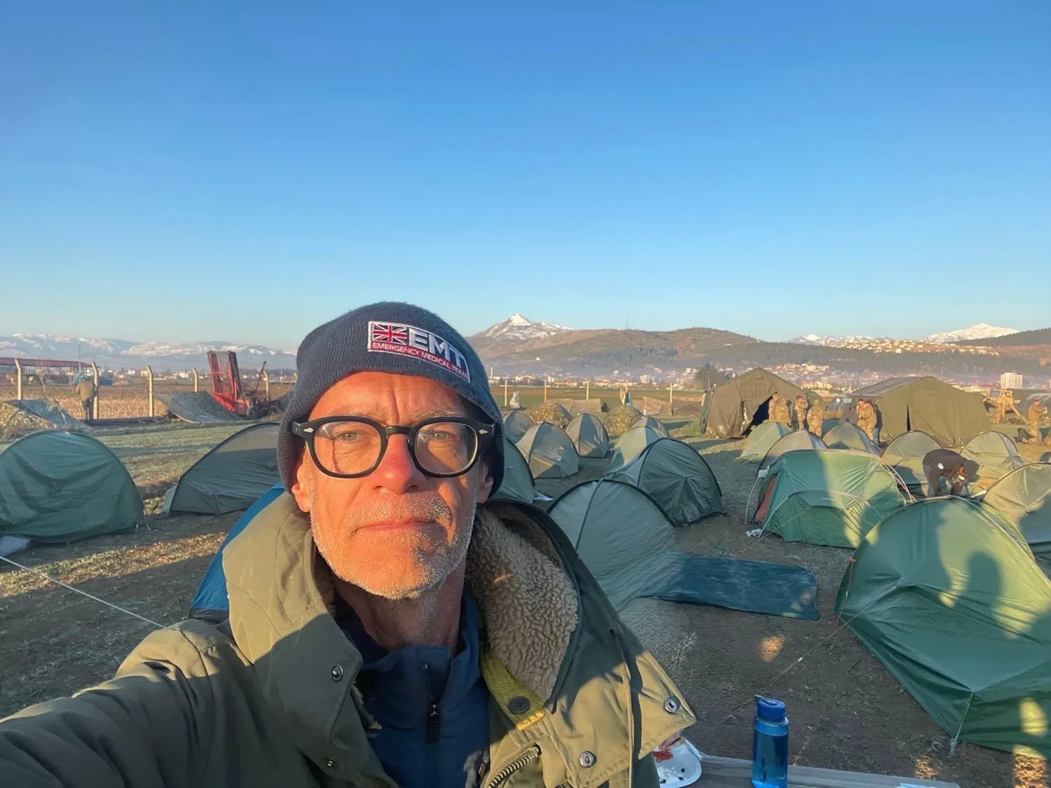 Johan von Schreeb in front of rows of green tents set up on dry ground with snow-capped mountains in the background under a clear blue sky.