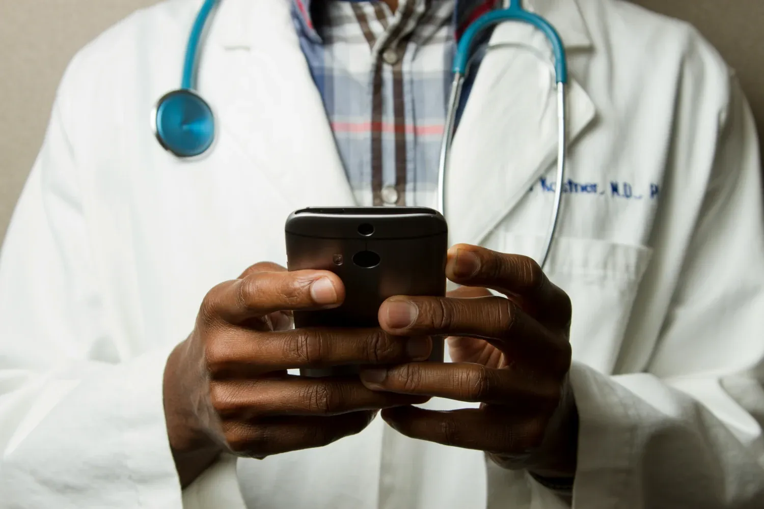 A person in a doctor's coat with a stethoscope around his neck is holding a smartphone.