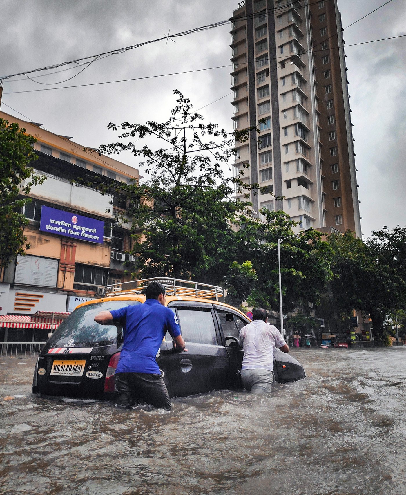 A street during a flood. Two people try to push a car through large masses of water.