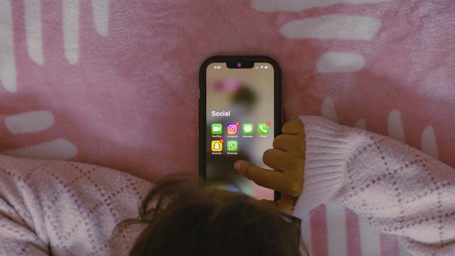 A child is lying in bed, using a smartphone that display various social media apps.