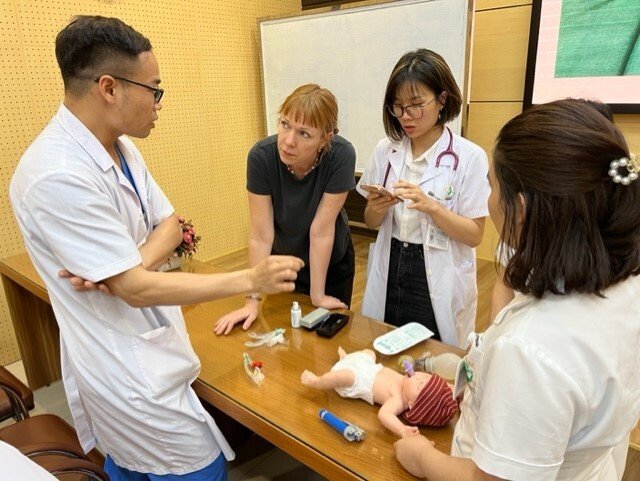 Susanna Myrnets Höök in the middle, leaning on a table discussing with three neonatologists during simulation trainings at Phu San Hospital. baby doll on the table for the simulation