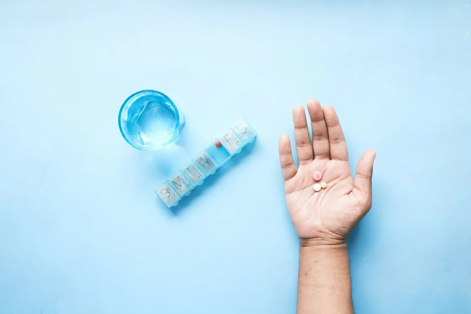 Genre image with blue background of a hand with pills, a pill box and a glass of water.