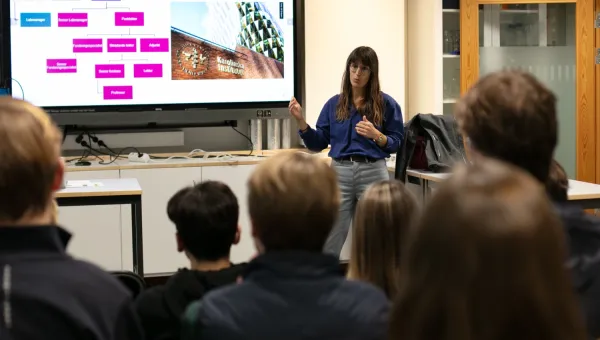 A woman stands in front of an audience in a classroom and gives a powerpoint presentation.