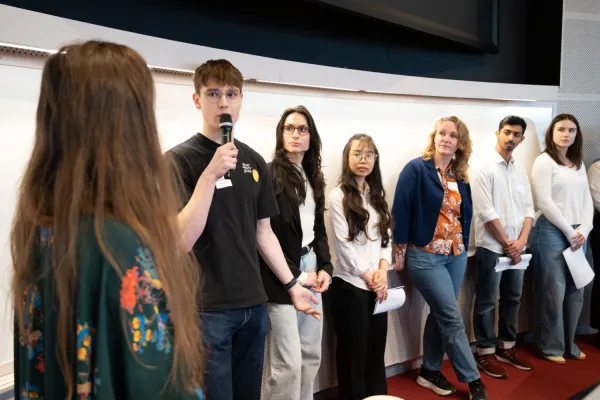 Students stand in a lecture hall. One person speaks into a microphone while other participants stand beside them holding papers.