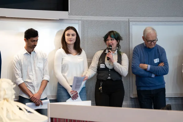 Students stand in a lecture hall. One person speaks into a microphone while other participants stand beside them holding papers.