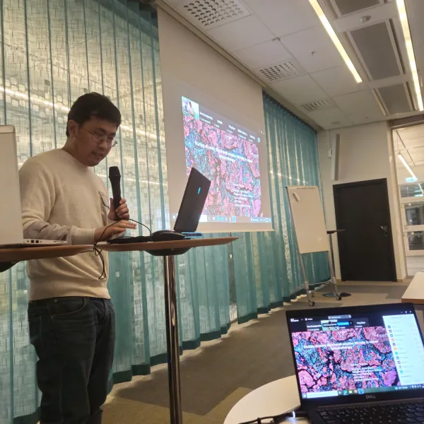 Photo of Yizhe Sun standing with a microphone in front of an audience in a larger meeting room