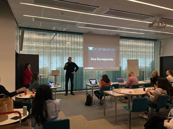 Photo of a person standing with a microphone in front of an audience in a larger meeting room