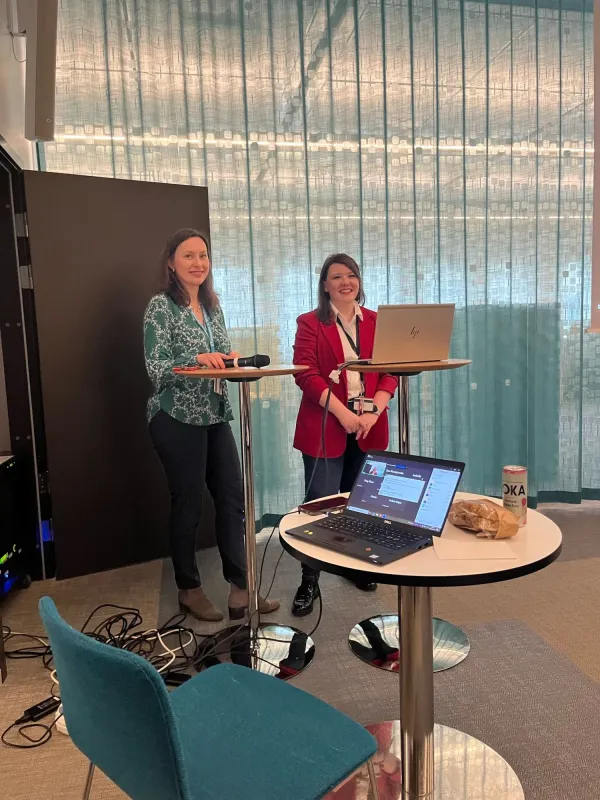 Photo of Tímea Szekerczés and Ewa Kurzejamska standing with a microphone in front of an audience in a larger meeting room