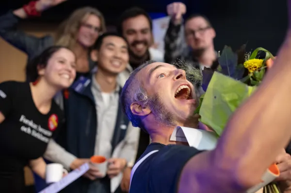 Happy man taking a selfie together with five people in the background whose faces are a bit blurred.