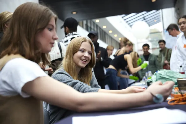 Two girls have put their arms on a table. In the background, high school students and researchers with white coats can be seen.