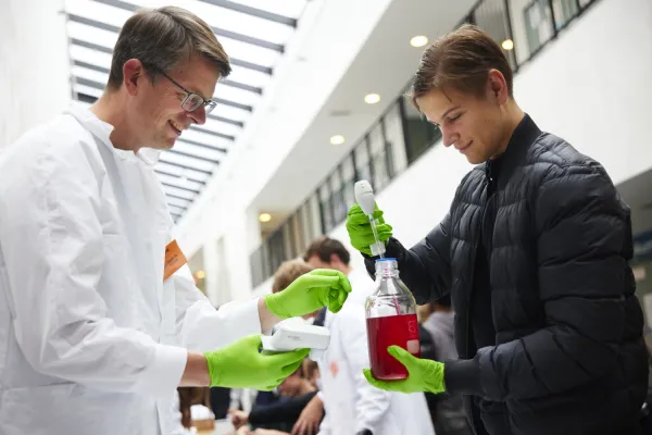 Researcher in white coat and green plastic gloves stands next to student with green plastic gloves. The student is holding a bottle of red liquid.