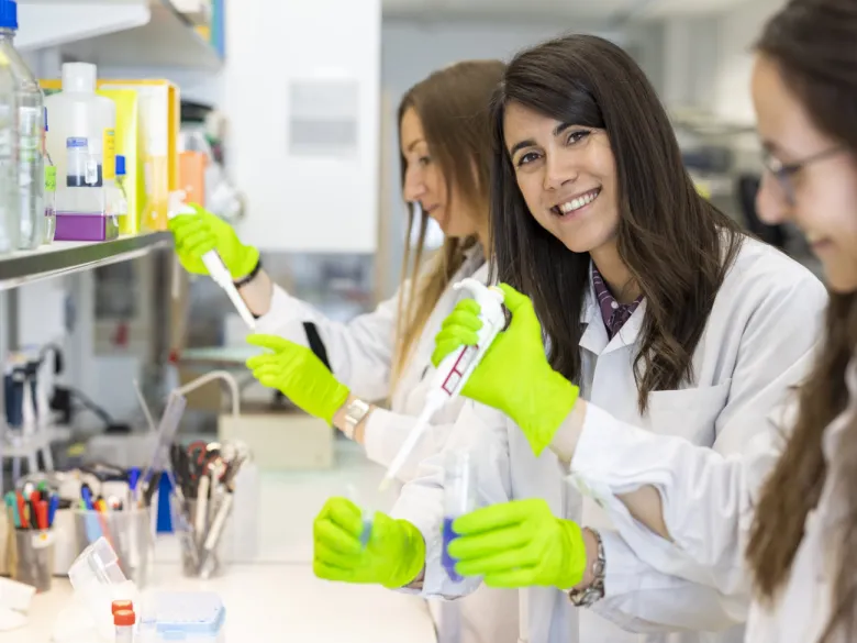 Three femare researchers standing with test tubes and pippettes in lab environment. One of them is looking in to the camera.