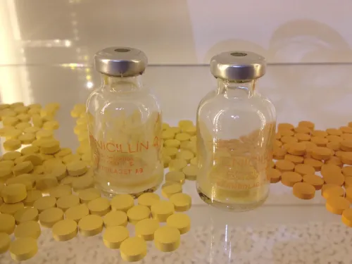 Two pill jar made of glass with the word penicillin on it, surrounded by yellow pills on a table