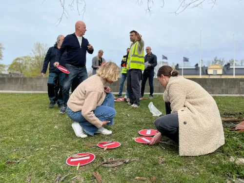 A group of people are standing in a grassy patch, looking at small round discs. One person is wearing a high-visibility vest.