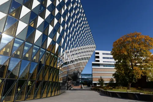 The glass facade of the Aula Medica. Next to it stands a large tree with colorful leaves.