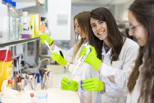 Three femare researchers standing with test tubes and pippettes in lab environment. One of them is looking in to the camera.