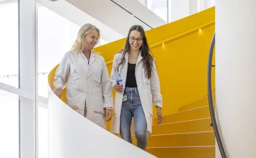 Two women in lab coats walking down yellow stairs.