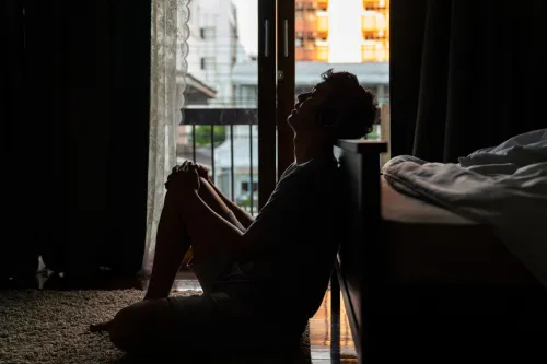 Depressed man sitting on the floor by a bed in a dark room.