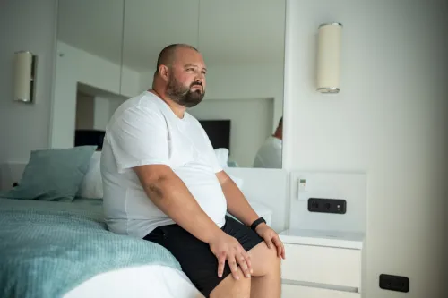 An overweight man is sitting on the edge of the bed in a brightly lit bedroom. He looks pensive.