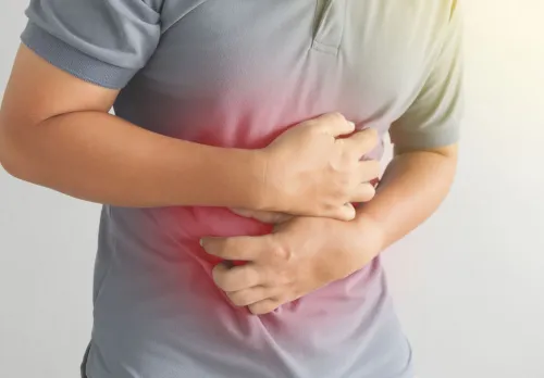 The image shows a person holding their stomach with both hands. The person is wearing a grey t-shirt and appears to be experiencing discomfort or pain in the abdominal area, indicated by a red light around the stomach.