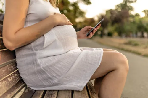 Side view of Young Unrecognizable pregnant , Caucasian woman sitting on a bench on a sunny day and using her smart phone while holding her tummy. Girl with mobile phone writing text on a brown bench in the park.