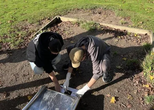 Two schoolchildren are crouching in front of a slide in a sandpit, collecting sand in a test tube.