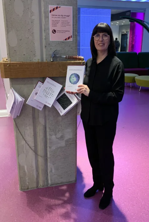 Woman standing with her thesis in hand in front of a pillar where several theses are nailed.