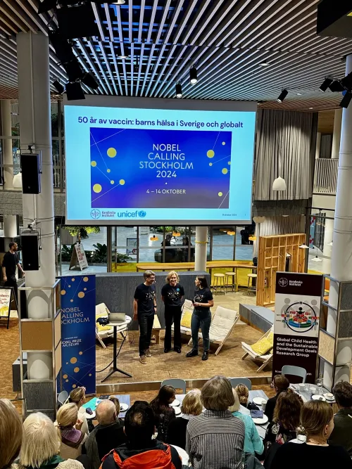 A group of three speakers stands on a stage in front of an audience, with a large screen displaying &quot;NOBEL CALLING STOCKHOLM 2024&quot; and event details. The setting is a modern venue with wooden accents and seating arranged for attendees. The speakers are di