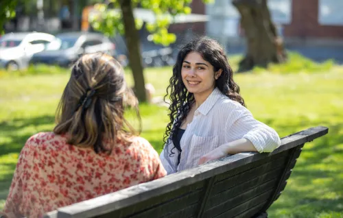 Students outside at campus Solna during a sunny spring day in 2022. || Two students sitting in the sun on a bench on campus. One looks into the camera and smiles.