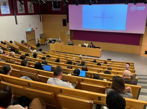 A lecture theatre with people watching four people and a PowerPoint presentation