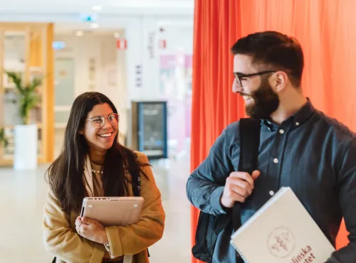 Two students laughing on campus. One of them is holding a laptop, the other a notebook with the logo of Karolinska Institutet.