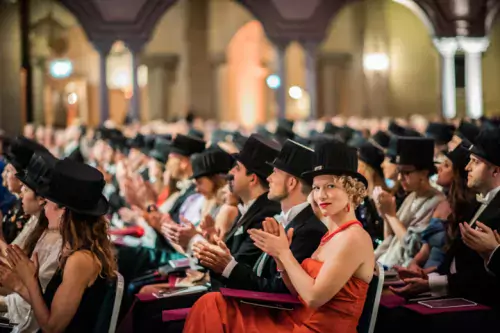 Newly minted doctors in their hats sit in the audience. One of them looks behind the camera.