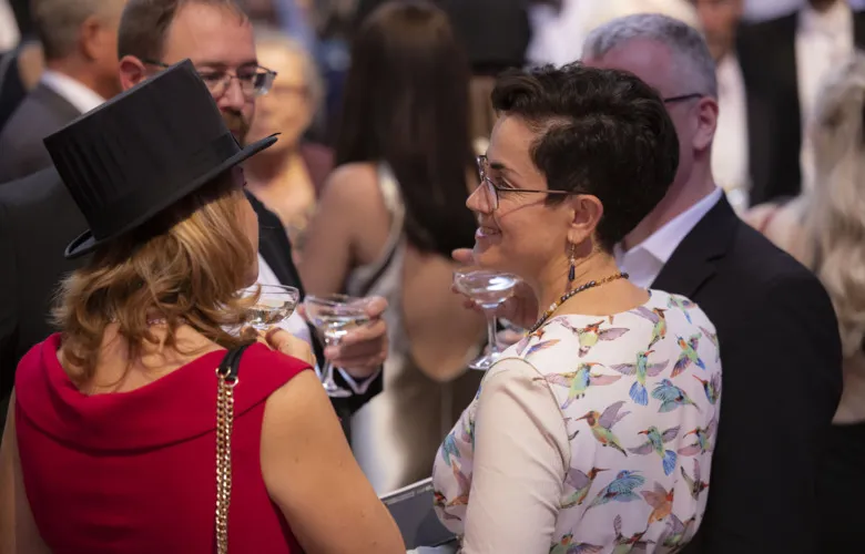 Two women, one of whom wears a doctor&#039;s hat, stand in a crowd and talk.